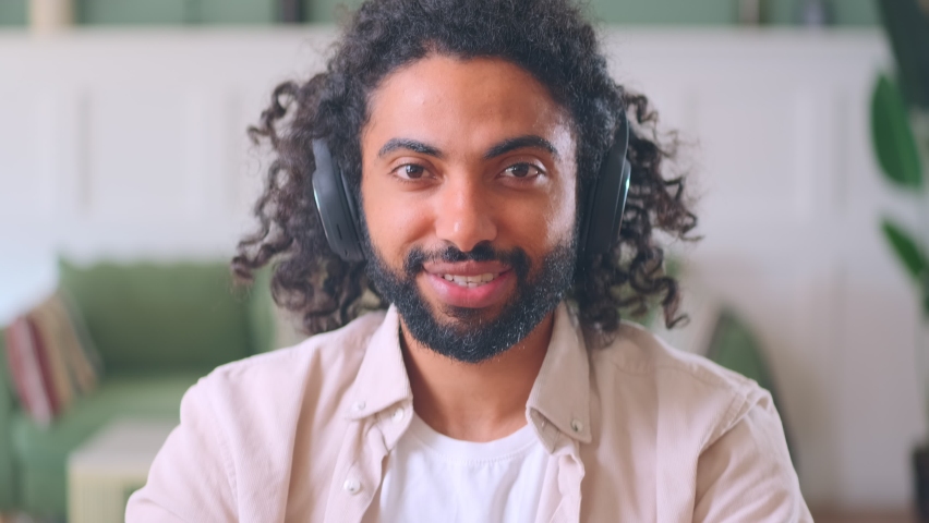Young friendly emotional middle eastern man wearing headphones, waving hand greeting interlocutor begins to say something and nodding talking with friends via video link sits in own house. camera view Royalty-Free Stock Footage #1092725639
