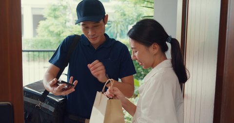 Young Asian delivery man in blue navy uniform knock door home delivery grocery online shopping to woman customer in front of the door and woman accept paper bag at house. Delivery service concept.