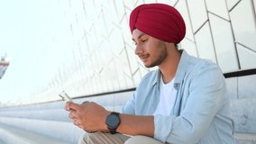Video call concept. Hilarious Indian man in casual shirt and traditional headwear turban using smartphone for virtual meeting, enjoys online chatting sitting on the steps in downtown, waving at