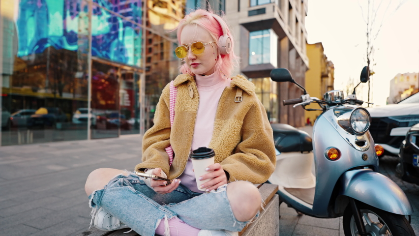 Coffee break in city center. Young positive lady with pink hair resting on bench and listening to music, web surfing on smartphone outdoors, tracking shot