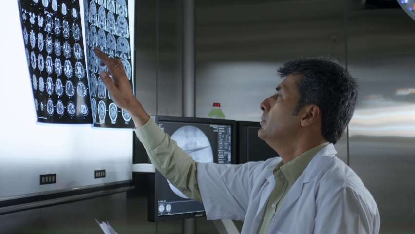 Middle aged Asian Indian male doctor, physician, or surgeon wearing a white apron or lab coat busy examining the X-Ray report and medical record standing in a hospital laboratory or operation theatre