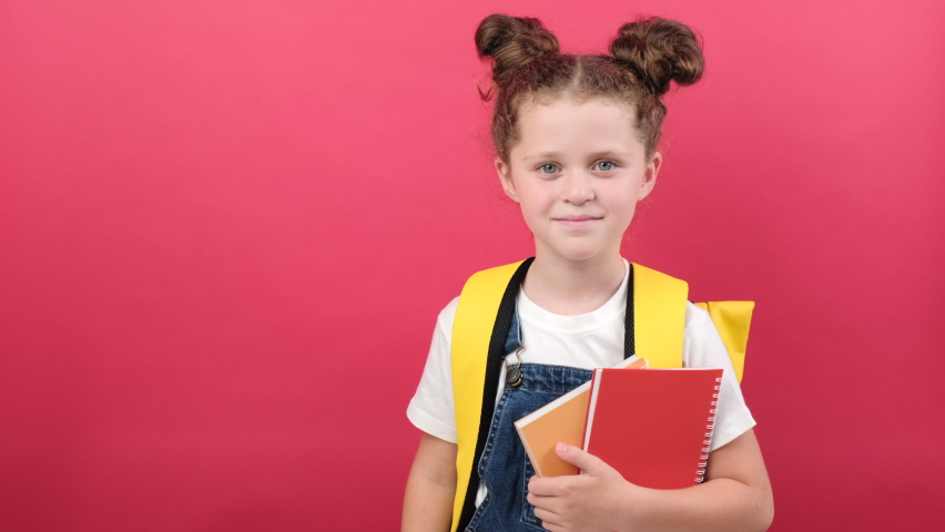 Portrait of school kid girl in white t-shirt and yellow backpack holding two notebooks, show ok okay hand gesture, posing isolated over red color background wall in studio. Kids education concept Royalty-Free Stock Footage #1092752101