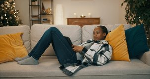 Beautiful girl of African appearance lies on couch in the living room. She is holding tablet and watching melodrama. The film moved the girl and she wiped away tear. A Christmas tree flickers behind.