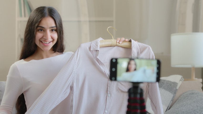 Young arabic beautiful blogger sits front of mobile phone on tripod records fashion clothing video for social network online translation shows shirt at camera enjoys clothes from online delivery Royalty-Free Stock Footage #1092754633