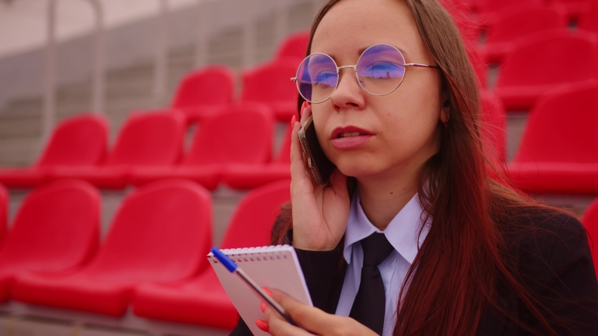 Young woman in glasses with notepad, pen talking on mobile phone, sitting on stadium bleachers.