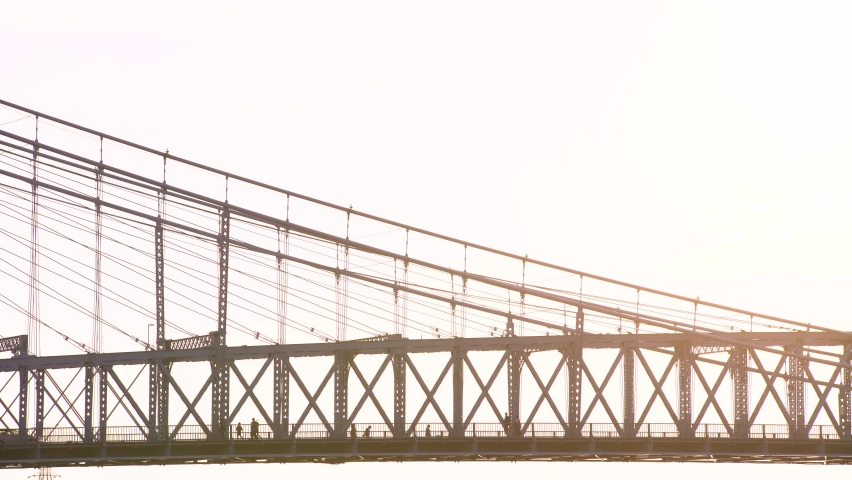 Timelapse of people crossing a suspension bridge at sunset. Silhouette profile shot of bridge with sun flare on bridge.