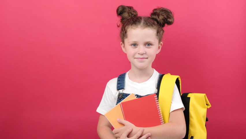 Portrait of school kid girl 6-7 years old in white t-shirt and yellow backpack holding two notebooks pointing fingers on workspace isolated over red background studio. Education lifestyle concept Royalty-Free Stock Footage #1092775123