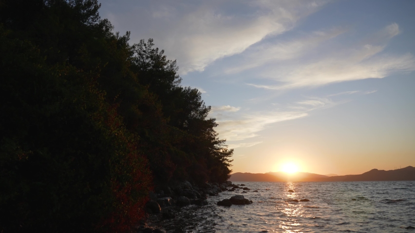 Sunset marine landscape. 4k stock video footage of beautiful peaceful dark evening sea water splashing, silhouettes of green trees, black mountains on horizon line, blue sky with white clouds and sun