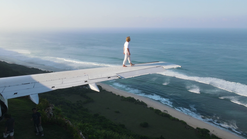 Handsome young man enjoys the stunning ocean view from the wing of an abandoned plane on a cliff in Bali
