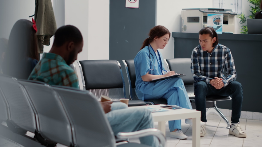Medical assistant filling in checkup report and talking to asian patient in hospital reception lobby. Nurse discussing about disease treatment and medicine support with person in waiting area. Royalty-Free Stock Footage #1092789673