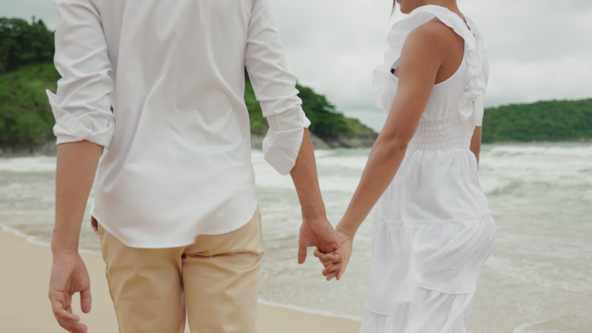 Rear view of a romantic couple holding hands and walking along a sandy beach. Bride and groom in casual white outfits enjoying a honeymoon vacation by the sea.