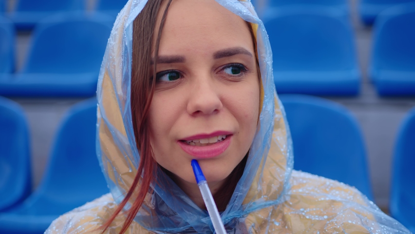 Young woman in raincoat with notepad, pen sitting on stadium bleachers alone in rainy weather. Female journalist writing down notes during sports training at street stadium.