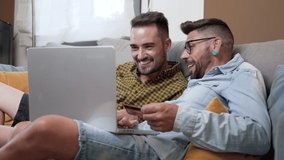 Happy young gay couple using laptop computer while sitting on a couch at home, shopping online with a credit card. High quality 4k footage