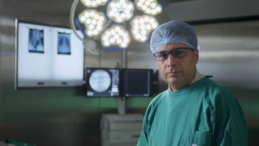 A middle-aged confident Indian Asian male surgeon, physician, or doctor wearing surgical apron, eyeglasses looking at the camera standing in an operation theatre. healthcare, medical, surgery concept 