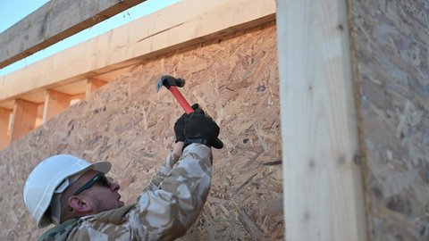 Carpenter hammering nail into OSB panel on the wall of future cottage. Close up of man worker building wooden frame house. Carpentry and construction concept.