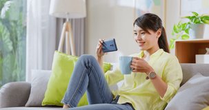 Asian woman sitting on chair using smartphone watch video or drama and drink beverage with mug feeling happy smiling when relax in living room at home