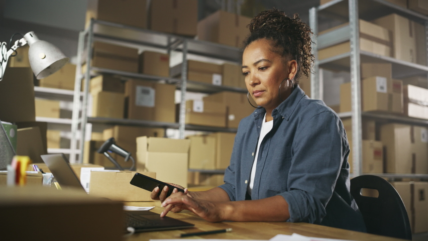 Inventory Manager Using Smartphone to Scan a Barcode on Parcel, Preparing a Small Cardboard Box for Postage. Black Multiethnic Small Business Owner Working on Laptop in Warehouse. Royalty-Free Stock Footage #1092830179