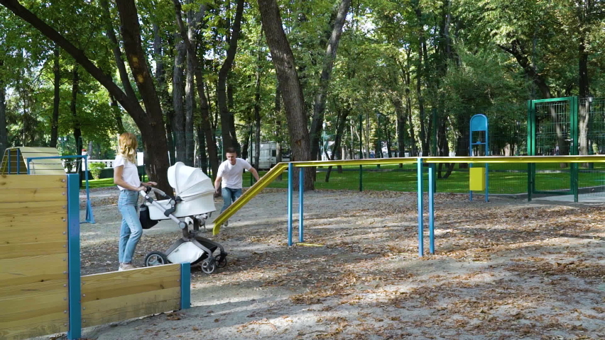 Jovial young man training Jack Russell Terrier on dog playground on summer day, blonde girl standing beside with baby stroller and watching them. Active family spending leisure together outside