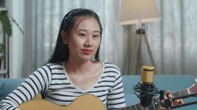 Close Up Of Asian Woman Waving Hand And Speaking To Camera While Live Stream Playing A Guitar At Home
