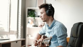 Happy millennial male artist in headphones and guitar have online video music lesson on computer. Practicing guitar in living room.
