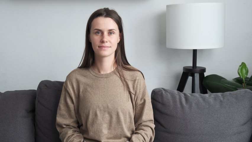 Sincere beautiful millennial young caucasian woman looking at camera, showing heart symbol, expressing love, feeling affection. Candid female volunteer demonstrating donation sign sitting on sofa