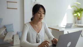 Cheerful Asian woman waves hello on video call with friend via modern laptop. Young employee takes break at work for online communication closeup