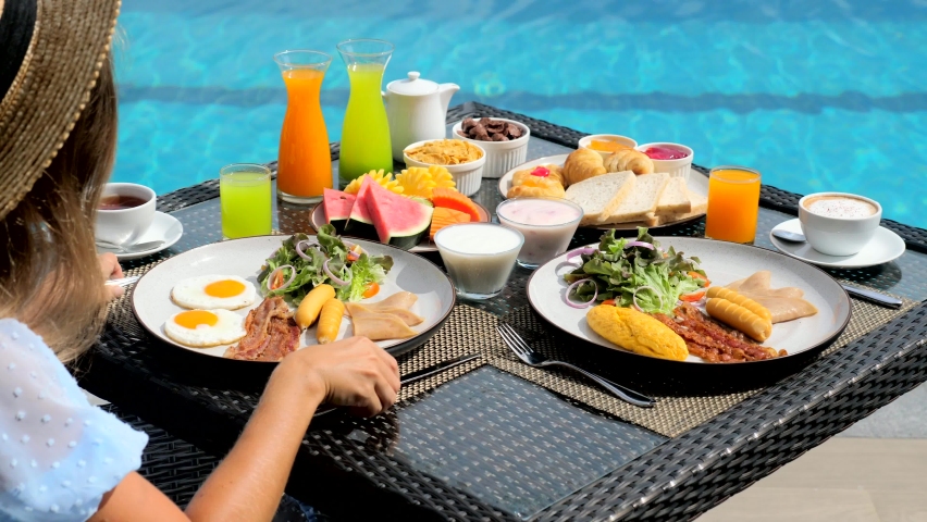Buffet service. Tasty breakfast served on table. Travel woman in hat eating breakfast is served with eggs, sausage, coffee, fresh orange juice, croissants, exotic fruits. Balanced diet on vacation | Shutterstock HD Video #1092842081