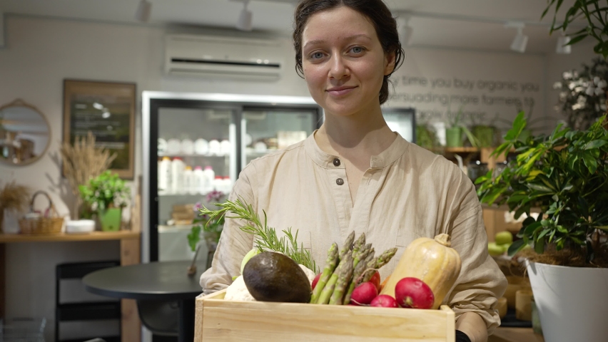 Woman with blue eyes looks at camera and smiles holding wooden box with fresh vegetables. Young woman with plait works in organic products shop