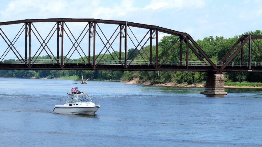 White boat with American flag approaches on the Mississippi River near a railroad bridge, at La Crosse, Wisconsin, on a beautiful day. Handheld clip.