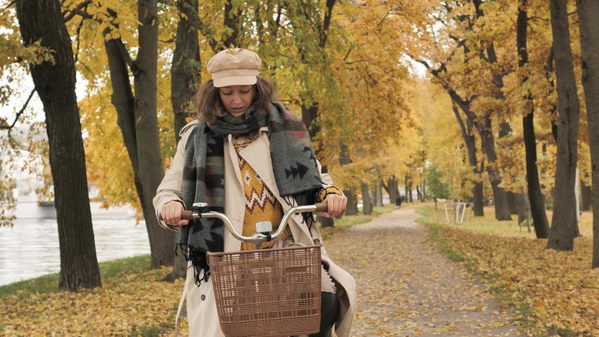 Woman dressed in trench coat scarf and cap rides bicycle between rows of trees on autumn park alley with fallen leaves closeup
