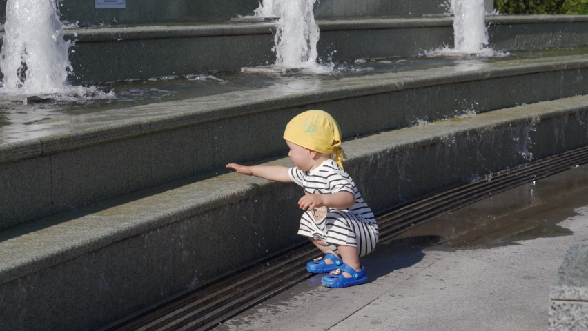 Happy kid having fun in fountain of public park at sunny summer day. 1.5 year old child playing with fountain water on a hot day.