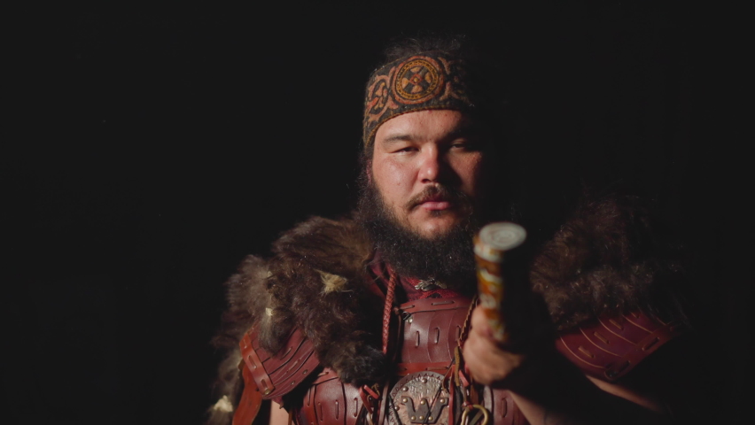 Obese man in ethnic leather costume with fur smokes shaman pipe on dark background slow motion. Folk magic rituals and worship. Hallucinogen tobacco