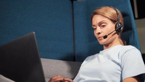 Confident middle aged pretty businesswoman wearing headset with mic sitting in coworking center enjoying video online conference call with foreign partners indoor.
