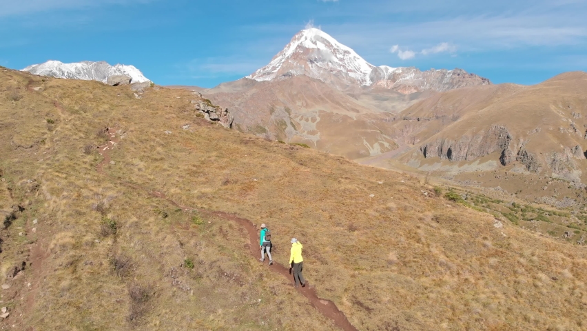 Two unidentified tourists with backpacks on the trekking in Caucasus mountains in Georgia. Aerial drone view of the young couple hiking with the majestic Kazbegi mountain at background, Georgia.