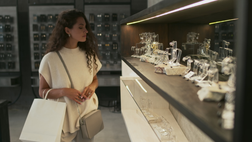 Medium long slow motion of young Biracial woman with shopping bags picking jewelry in modern luxury shop, smiling Royalty-Free Stock Footage #1092861601
