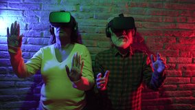 multiracial senior couple play with virtual reality goggles in a brick-walled house, asian man with african american woman.