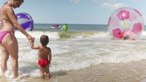 A little cute cheerful kid, looks at horizon of blue calm sea and bright inflatable big balls for children with joyful kind happy mother in bright bikini under the scorching sun. 4K UHD slow-mo
