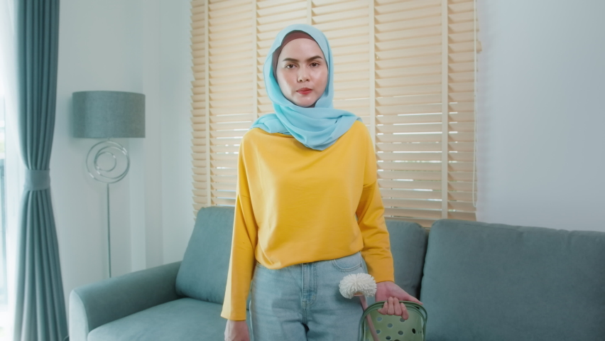 Young happy muslim woman wearing yellow gloves and holding a basket of cleaning supplies in living room.