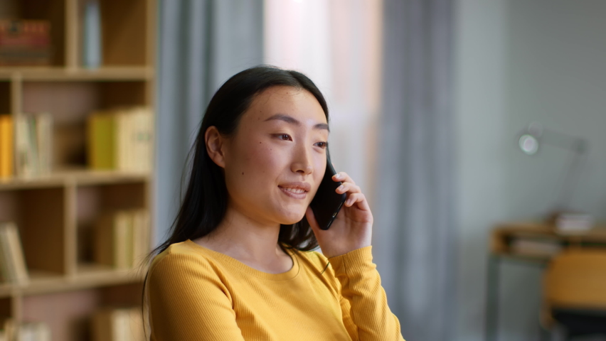 Mobile communication. Close up portrait of young happy carefree asian woman talking on cellphone with friends, enjoying pleasure talk at home, slow motion, free space