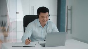Corporate virtual meeting. Happy middle aged asian man employee in headset video chatting with colleagues online via laptop, taking notes in office and laughing, slow motion