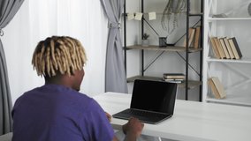 Online meeting. Casual man. Virtual communication. Hipster black guy sitting home desk talking to opened laptop blank screen in light room interior.
