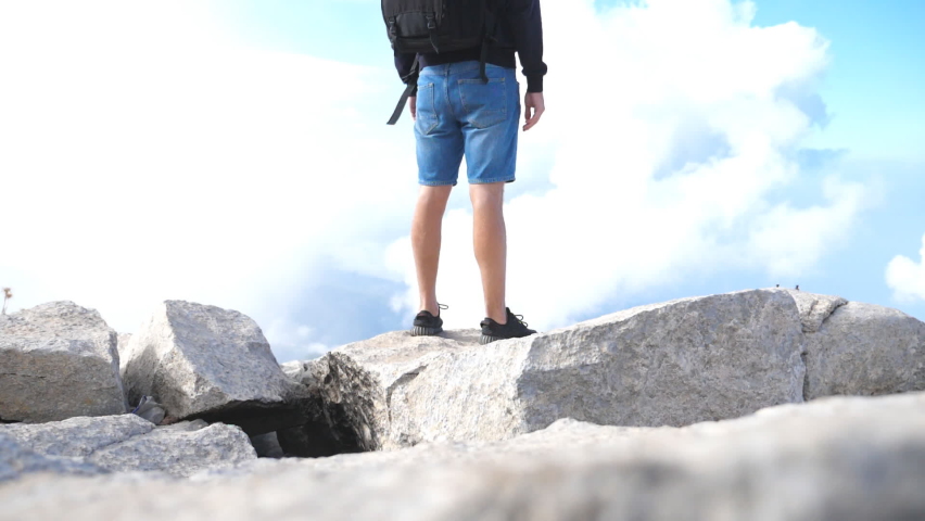 Boy tourist with backpack standing on the peak of the hill and victoriously raising hands. Young male hiker reaching up top of mountain and outstretching arm. Man enjoying freedom during summer travel