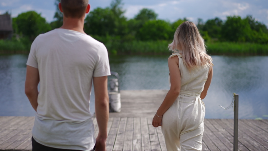Tracking shot of excited young couple running in slow motion to river on wooden pier smiling. Back view Caucasian man and woman having fun enjoying leisure at eco-resort outdoors. Nature tranquility