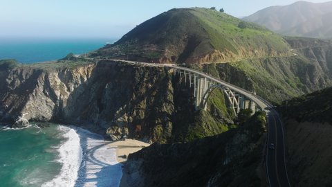 Cinematic aerial shot SUV cars driving on sunny clean road highway to mountains. Summer vacation dream travel, road trip by cinematic nature landscape at Big Sur with Bixby arch bridge on West Coast