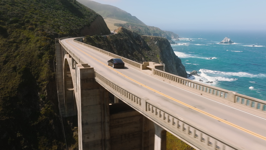 Electric car driving by scenic bridge on cinematic ocean background. Electric sedan drives by mountain coast road, scenic route. Alternative energy for cars. Eco and environmentally friendly Royalty-Free Stock Footage #1092882799