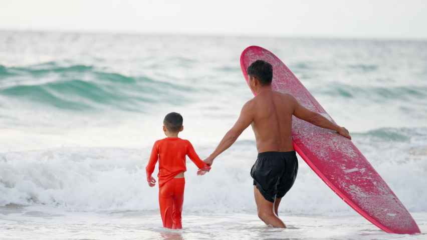 Happy Asian family on summer vacation. Grandfather teaching grandchild boy surfing on surfboard in the sea. Senior man and little boy kid enjoy outdoor activity lifestyle water sport surfing together.