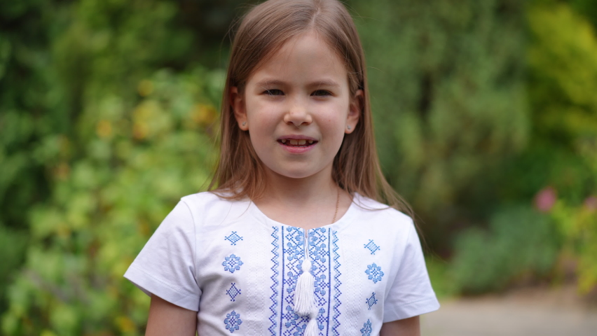 Brunette charming Ukrainian girl in national shirt drawing colors of flag on cheek looking away talking. Portrait of pretty child posing in summer garden outdoors. Childhood and Ukraine concept
