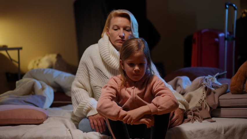 Portrait of stressed Ukrainian little daughter sitting in bomb shelter on air-raid alert warning as mother closing child ears with hands talking. Anxious tired girl and woman hiding from bombardment