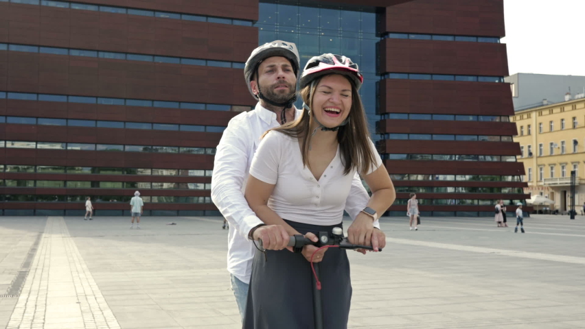 Young man rides his girlfriend on an electric scooter around the city square. Both smile happily.