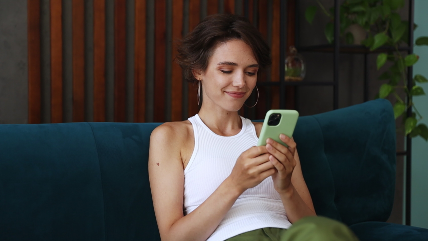 Young woman wears casual clothes sits on blue sofa hold using mobile cell phone found out great big win news do winner gesture say yes stay home rest relax spend time in living room indoors grey wall
